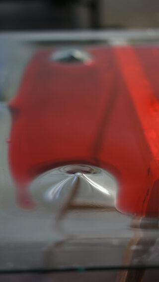 Nahaufnahme einer roten Flüssigkeit, die über eine flache Oberfläche gegossen oder verteilt wird, wodurch eine verzerrte Reflexion und ein glänzender, erhabener zentraler Bereich entstehen.