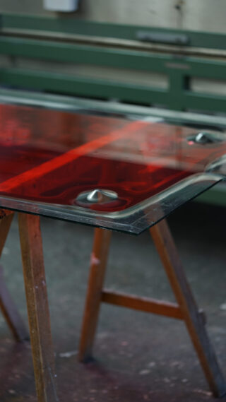 Eine rechteckige Glasscheibe mit roten Reflexen liegt auf zwei hölzernen Sägeböcken, auf deren Oberfläche Metallgegenstände platziert sind, in einer industriellen Umgebung.