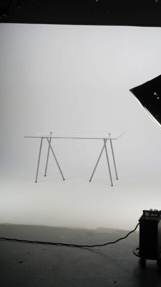 Ein Tisch mit Glasplatte und Metallbeinen steht in einem Fotostudio mit weißem Hintergrund und sichtbarer Beleuchtungsausrüstung.