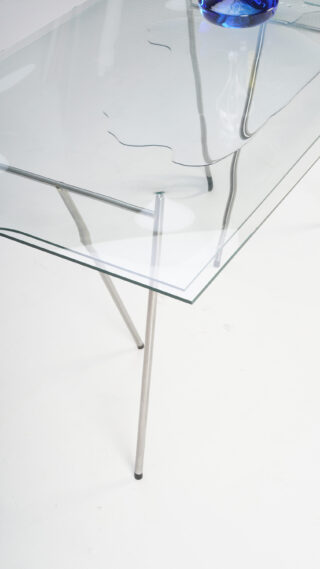 Ein Glastisch mit einem großen, unregelmäßigen Riss auf der Oberfläche; Metallbeine und ein blaues Objekt spiegeln sich auf dem Tisch.