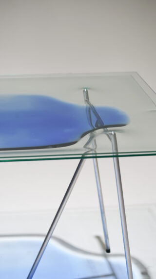 Eine Nahaufnahme eines Glastisches, auf dessen Oberfläche eine blaue Flüssigkeit verschüttet ist. Der Tisch wird von dünnen, abgewinkelten Metallbeinen vor einem schlichten Hintergrund getragen.