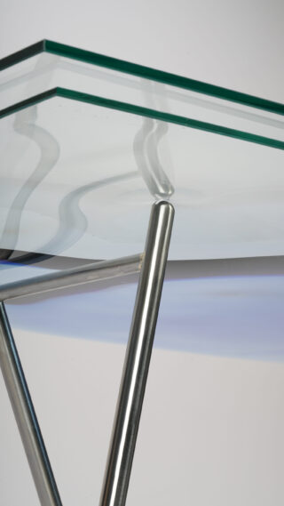 Eine Nahaufnahme einer Glastischplatte, die von abgewinkelten Metallbeinen getragen wird, wobei auf der Glasoberfläche Reflexionen sichtbar sind.