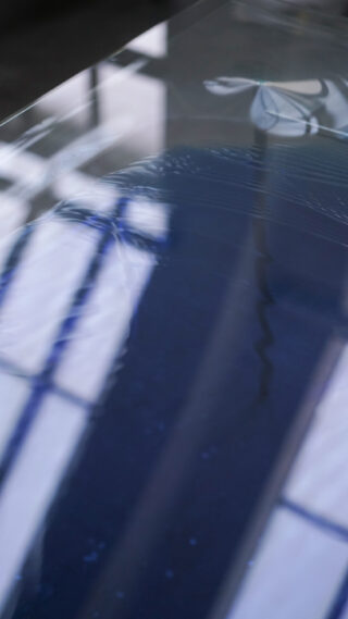 Eine Nahaufnahme einer reflektierenden Glasoberfläche, die teilweise Reflexionen von Fensterscheiben und ein verzerrtes Blumenmuster zeigt.