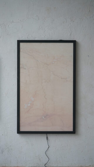 An einer grauen Wand hängt ein rechteckiger Rahmen mit marmorierter Oberfläche, von dem aus eine schwarze Kordel nach unten verläuft.