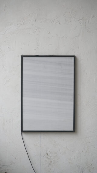 An einer strukturierten, cremefarbenen Wand hängt ein quadratisches elektronisches Panel mit dünnem Rahmen und sichtbarem Kabel.