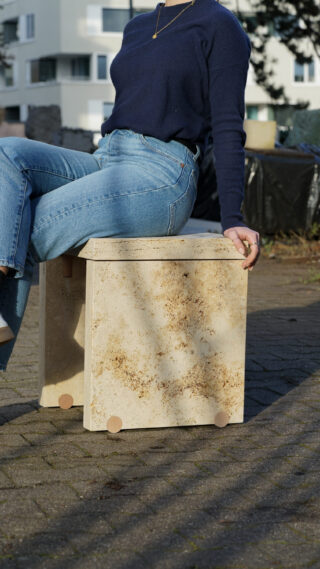 Eine Person in Blue Jeans und einem marineblauen Pullover sitzt im Freien auf einer beigen Steinkiste auf einer gepflasterten Fläche, im Hintergrund sind Gebäude und Grünflächen zu sehen.