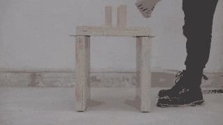 Eine Person demonstriert den Zusammenbau eines kleinen Holztisches, indem sie flache Platten zusammensteckt und zwei zylindrische Objekte darauf platziert.