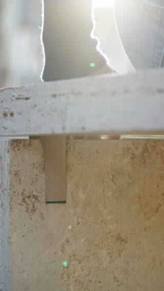 Nahaufnahme einer verwitterten Stein- oder Betonoberfläche mit grellem Sonnenlicht, einer stützenden Holzhalterung darunter und dem Fuß einer Person, der an der oberen Kante sichtbar ist.