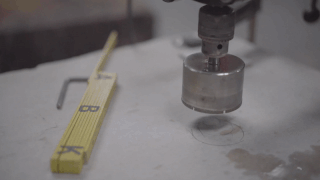 Ein an einer Bohrmaschine befestigter Lochsägenbohrer drückt nach unten und schneidet ein kreisförmiges Loch in eine flache, helle Oberfläche; daneben liegt ein gefaltetes Messlineal.