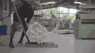 Eine Person verwendet einen langen Holzhebel, um einen großen, unbearbeiteten Steinblock in einer geräumigen Industriewerkstatt anzuheben und zu bewegen.