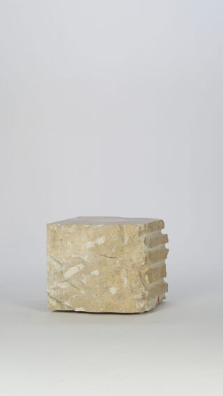 Ein etwa rechteckiger Block aus beigem Stein mit einer unbearbeiteten, strukturierten Oberfläche liegt auf einem schlichten, hellen Hintergrund.