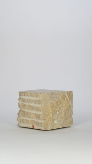Auf einem schlichten weißen Hintergrund befindet sich ein grob behauener, rechteckiger Block aus beigem Stein mit horizontalen Rillen auf einer Seite.