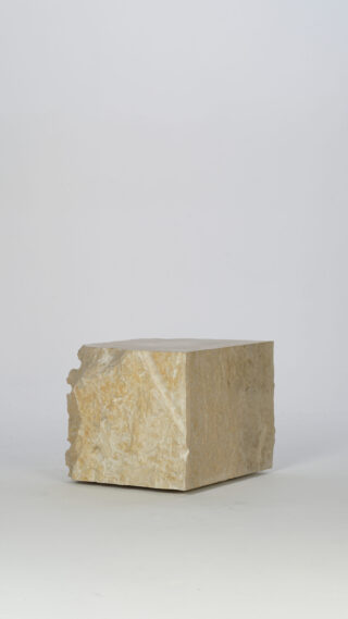 Ein rechteckiger Block aus rauem, hellem Stein liegt auf einer schlichten, weißen Oberfläche vor einem neutralen Hintergrund.