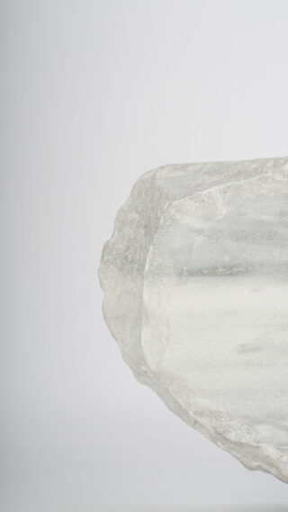 Nahaufnahme eines rauen, durchscheinenden weißen Minerals oder Kristalls vor einem schlichten hellgrauen Hintergrund, der die rechte Seite des Bildes einnimmt.