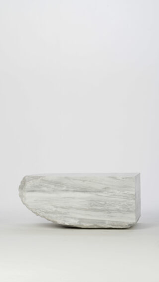 Rechteckiger Block aus gräulichem Stein mit rauer, unebener linker Kante, vor einem schlichten hellen Hintergrund.