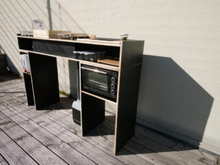 Eine minimalistische Außenküche mit Regalen, Toaster, Behältern, Wasserspender und Küchenutensilien auf einem Holzdeck neben einer hellen Wand.