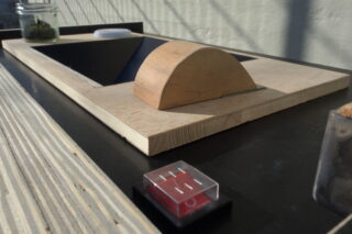 Eine Nahaufnahme eines kuppelförmigen Holzobjekts auf einem flachen Brett, im Hintergrund ein Glasgefäß und im Vordergrund eine kleine durchsichtige Schachtel mit roten Fächern.