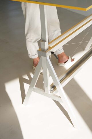 Eine Person in weißen Hosen und Sandalen steht neben einem weißen Metalltisch mit gelben Kanten und wirft Schatten auf einen hellen Boden.