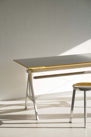 Ein minimalistischer Arbeitsbereich mit einem Tisch mit Metallrahmen und einem dazu passenden Hocker, beide mit gelben Akzenten, im Sonnenlicht vor einer schlichten weißen Wand positioniert.