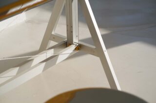 Nahaufnahme einer weißen Holzstaffelei mit einem Metallscharnier und einer daran befestigten Schnur, die auf einem hellen Boden im direkten Sonnenlicht steht.