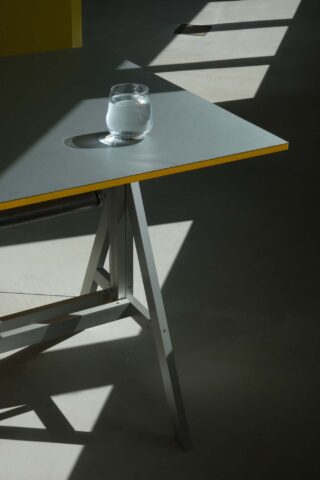 Auf der Ecke eines grauen Tisches mit Metallbeinen steht ein Glas Wasser. Im Sonnenlicht, das durch ein Fenster auf den Boden fällt, wirft es einen Schatten.
