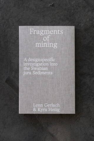 Auf einer dunklen Steinfläche liegt ein graues Buch mit dem Titel „Fragmente des Bergbaus: Eine gestaltungsspezifische Untersuchung der schwäbischen Alb-Sedimente“ von Lenn Gerlach und Kyra Heilig.