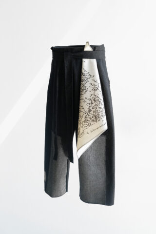 Ein schwarzer asymmetrischer Rock mit mehrlagigen Einsätzen und einem weißen Stoffeinsatz mit einem schwarz gedruckten Muster auf einem schlichten weißen Hintergrund.