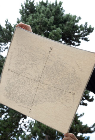 Person, die vor einem grünen Baum und bewölktem Himmel ein Stoffquadrat mit einer Kompassrose und schwachen Kartenmarkierungen hochhält.