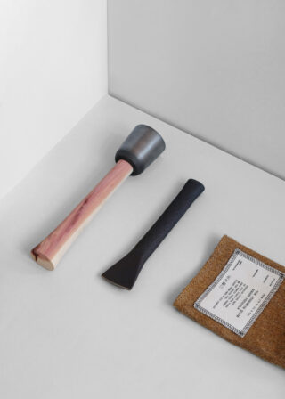 Ein Hammer mit Holzgriff, ein schwarzer Metallmeißel und ein brauner rechteckiger Beutel mit weißem Etikett sind auf einer weiß-grauen Oberfläche angeordnet.