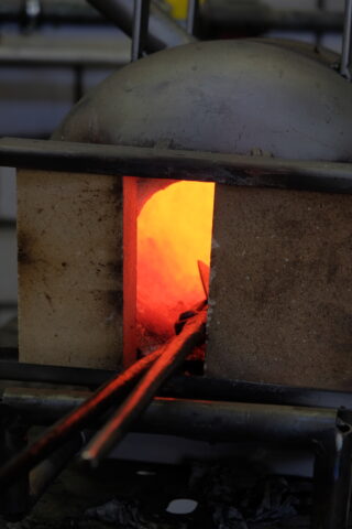 In einem kleinen Industrieofen wird ein Metallstab erhitzt. Durch die Ofenöffnung ist ein hell orangefarbenes Glühen sichtbar.