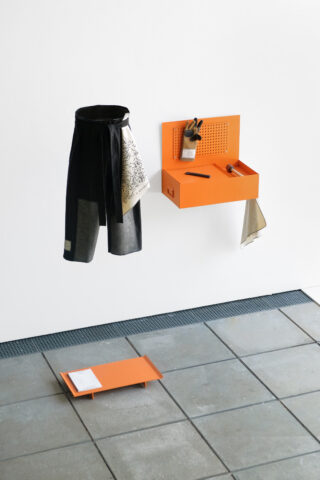 Ein orangefarbener Wand-Organizer mit Fächern und Haken dient zur Aufbewahrung von Utensilien und Stoffen; darunter steht auf einem gefliesten Boden neben einer hängenden schwarzen Schürze ein passendes Tablett.