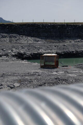 Ein Metallkäfig mit Ausrüstung darin steht auf felsigem Boden in der Nähe eines Gewässers, im Vordergrund ist ein großes gewelltes Metallrohr zu sehen.
