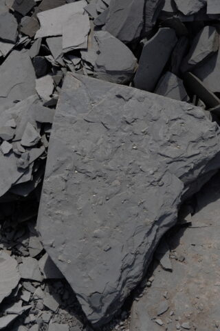 Ein großer, flacher, grauer Stein mit rauen Kanten liegt zwischen kleineren, zerbrochenen Stücken ähnlicher grauer Steine.