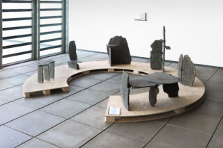 Auf einer halbrunden Holzplattform sind in einem minimalistischen, gefliesten Raum aufrechte Platten aus dunklem Stein zu sehen, die in verschiedenen abstrakten Formationen angeordnet sind.