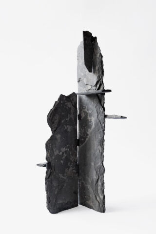 Eine vertikale skulpturale Struktur aus grob strukturierten, dunklen, schieferartigen Materialien, die sich im rechten Winkel kreuzen und vor einem schlichten weißen Hintergrund stehen.