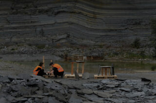 Zwei Personen in orangefarbenen Sicherheitswesten sitzen und untersuchen Steine in einem Steinbruch, umgeben von Schieferstücken und geschichteten Felsformationen.