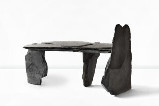 Ein Tisch aus unregelmäßig geformten Schieferplatten mit drei Beinen aus grobem Stein und einer flachen Schieferplatte vor einem schlichten weißen Hintergrund.