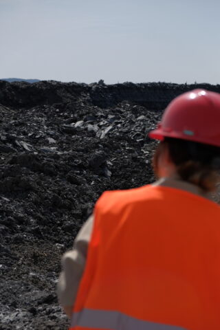 Eine Person in einer orangefarbenen Sicherheitsweste und einem rosa Schutzhelm steht unter klarem Himmel vor einer großen, felsigen Industriegrube.