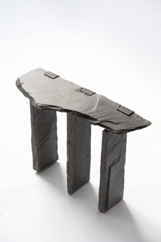 Ein minimalistischer Tisch aus drei vertikalen Schieferplatten, die eine einzelne unregelmäßig geformte Schiefertischplatte tragen, vor einem schlichten weißen Hintergrund.
