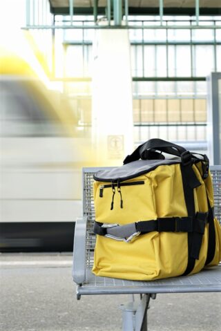 Ein gelber Rucksack liegt auf einer Metallbank an einem Bahnhof, im Hintergrund fährt unscharf ein Zug vorbei.