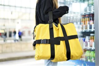 Eine Person in einer schwarzen Jacke hält eine große gelbe Reisetasche mit schwarzen Riemen und steht neben einem Getränkeautomaten.