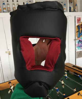 Eine Hand hält eine schwarz-rote gepolsterte Kopfbedeckung über einem Tischfußballtisch, im Hintergrund sind Flaschen, Schränke und Wandkunst zu sehen.