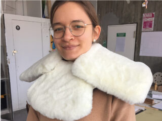 Eine Person mit Brille und braunem Mantel trägt in einer Inneneinrichtung mit Industriedekor einen großen, weißen Plüschkragen.