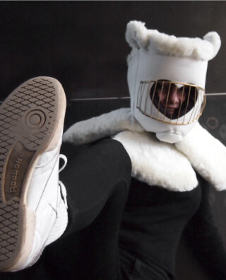 Person in einem weißen, bäreninspirierten Kostüm mit einem goldenen Käfig über dem Gesicht, schwarzer Kleidung und weißen Turnschuhen, sitzt und hebt einen Fuß in Richtung Kamera.