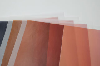 Mehrere durchscheinende Papierblätter in unterschiedlichen Braun-, Orange- und Pfirsichtönen sind in einem fächerartigen Muster auf einer weißen Oberfläche angeordnet.