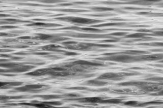 Nahaufnahme der Wasseroberfläche mit Wellen und Spiegelungen, dargestellt in Schwarzweiß.
