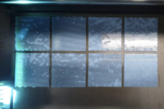 Acht rechteckige Abschnitte mit ungleichmäßiger transluzenter Beschichtung sind unter heller Beleuchtung in zwei Reihen auf einer metallischen Oberfläche angeordnet.