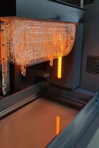 Ein 3D-Drucker im Betrieb mit einem teilweise gedruckten transparenten Objekt, das über einem Harzbehälter hängt und von einem orangefarbenen Licht beleuchtet wird.
