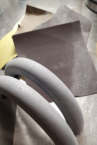 Zwei gebogene Metallobjekte mit sichtbaren Schleifspuren liegen auf einer metallischen Oberfläche neben Schleifpapierblättern in verschiedenen Farben.