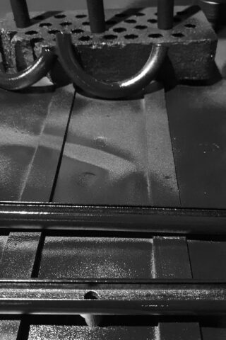 Schwarzweißbild, das Metallrohre zeigt, die mit einem rechteckigen Block mit Löchern verbunden sind, der auf einer flachen Oberfläche mit starken Schatten und Reflexionen ruht.